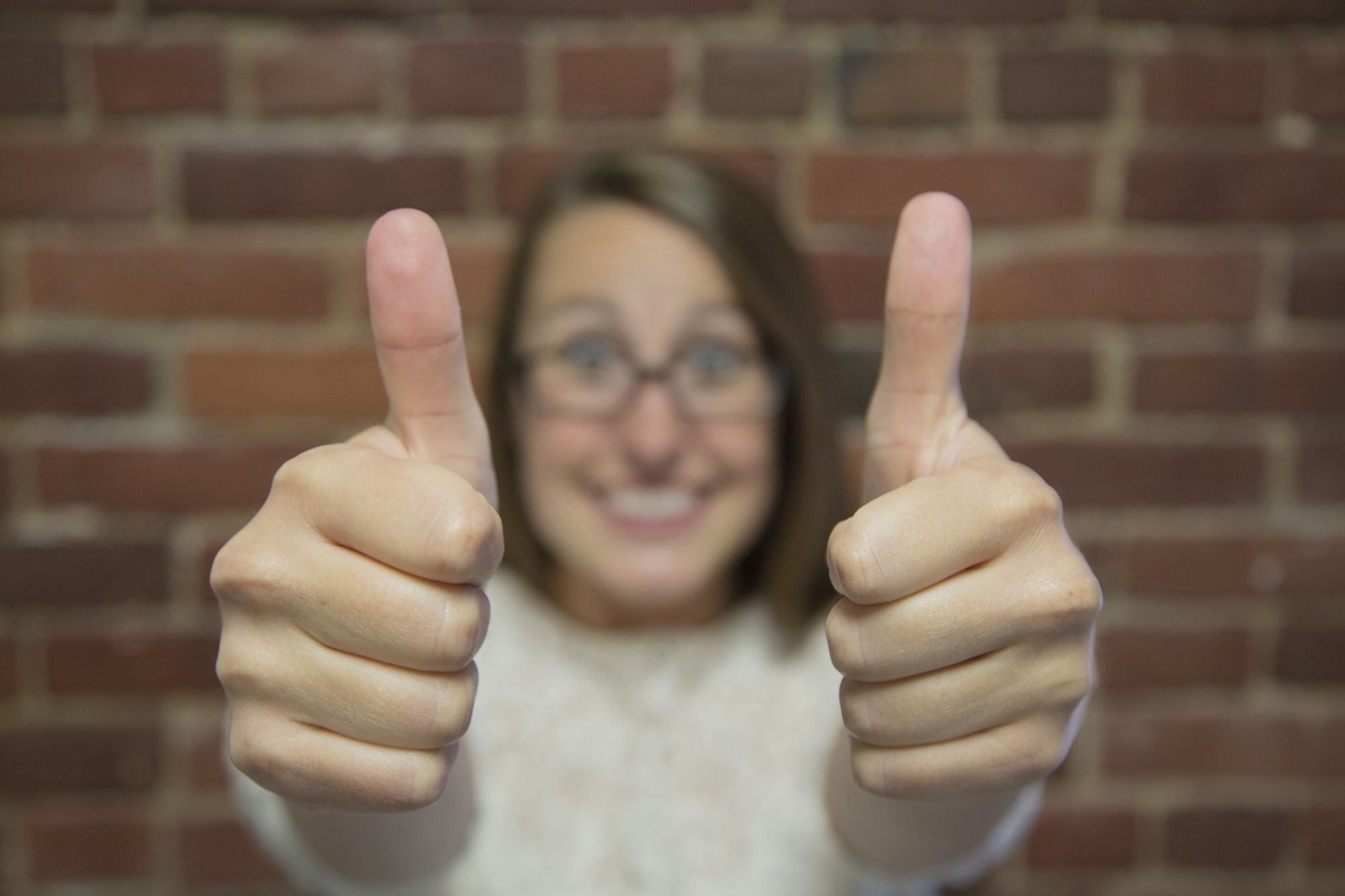 women in front of brick wall with two thumbs up smiling wearing glasses